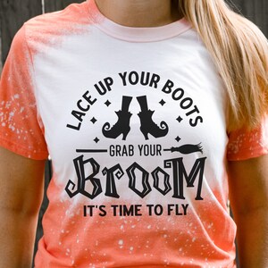 May include: A white t-shirt with an orange ombre design featuring a black graphic that says "Lace Up Your Boots Grab Your Broom It's Time To Fly" with a witch's hat and broom illustration.