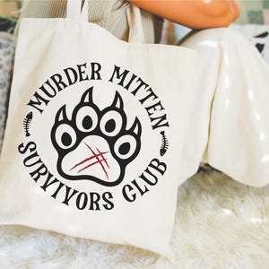 Pu&ograve; includere: Una borsa tote bianca con un design a forma di impronta di zampa nera e rossa. Il testo "Murder Mitten Survivors Club" &egrave; scritto intorno all'impronta di zampa.