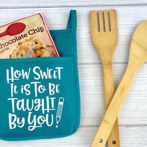 May include: Teal oven mitt with a pocket that has a package of chocolate chip cookie mix inside. The oven mitt has the text "How Sweet It Is To Be Taught By You!" with a pencil graphic.