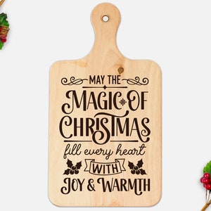 Puede incluir: Una tabla de cortar de madera con el texto "May the magic of Christmas fill every heart with joy & warmth" en una fuente marr&oacute;n.
