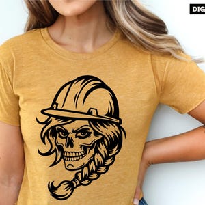 Op de afbeelding: Een zwart-witte illustratie van een schedel die een helm en een vlecht draagt. De schedel heeft een woeste uitdrukking en draagt een helm met een rand. De vlecht is lang en golvend. De illustratie is op een geel T-shirt.