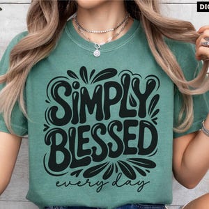 May include: A teal t-shirt with the words "SIMPLY BLESSED" in large, black, stylized letters, with "every day" below. The shirt has a relaxed fit and short sleeves. The words "DIGITAL DESIGN" are in the top right corner.