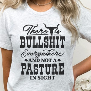 May include: A white t-shirt featuring a black graphic that reads "There is BULLSHIT Everywhere AND NOT A PASTURE IN SIGHT". The graphic also includes a stylised bull skull.