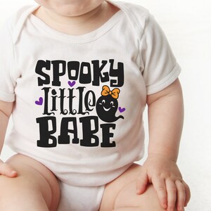 May include: A white baby onesie with the text "Spooky Little Babe" in black. A purple ghost with a pink bow is next to the text.