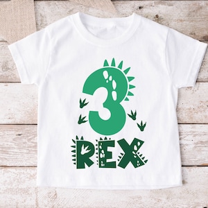 Op de afbeelding: Witte peuter-T-shirt met een groen dinosaurusontwerp. Het nummer "3" is gestileerd met een dinosaurusmotief, vergezeld van dinosaurusvoetstappen en het woord "REX". Het ontwerp is gecentreerd op het shirt.