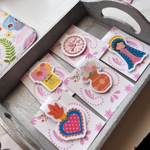 May include: A set of five colorful bookmark magnets with a pink and white background. The magnets feature a hummingbird, a vase of flowers, a heart, a religious figure, and a jar of salsa. The magnets are in a wooden tray.