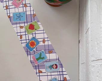 May include: A decorative strip featuring colorful illustrations of Mexican Loteria cards. The cards are set against a light purple and white checkered background, with brown bean-shaped accents. The strip is held near a clear container of colorful pens.
