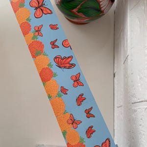 May include: A blue decorative paper strip featuring orange marigold flowers and red butterflies. The strip is being held near a white wall. A vase with flowers is in the background. A clear container with colorful pens is in the lower left corner.