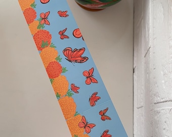 May include: A blue decorative paper strip featuring orange marigold flowers and red butterflies. The strip is being held near a white wall. A vase with flowers is in the background. A clear container with colorful pens is in the lower left corner.