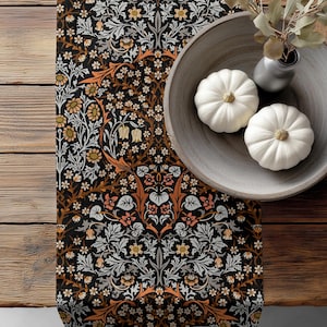 May include: A table runner with a black background and a floral pattern in orange, white, and gold. Two white pumpkins sit in a wooden bowl, next to a small vase with greenery. The runner is on a wooden table.
