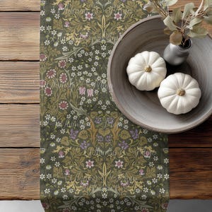 May include: A decorative table runner with a floral pattern in shades of green, gold, and purple. Two small white pumpkins sit in a wooden bowl, alongside a small vase with greenery. The runner is on a wooden table.