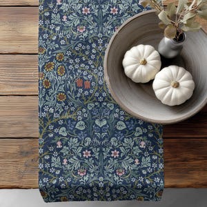 May include: A navy blue floral table runner with a detailed pattern of flowers and leaves. Two white pumpkins sit in a wooden bowl, next to a small vase with greenery. The runner is on a wooden table.