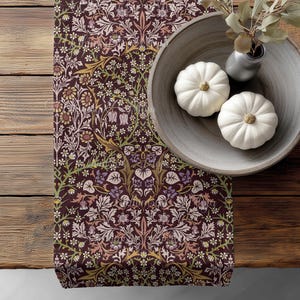 May include: A decorative table runner with a floral pattern in shades of burgundy, gold, and purple. Two small white pumpkins sit in a wooden bowl, next to a small vase with greenery. The runner is on a wooden table.