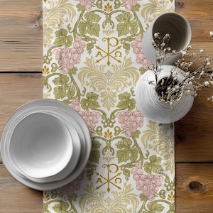 May include: A white table runner with a repeating pattern of pink grapes, green leaves, and gold accents. The runner also features gold Chi Rho symbols. Two white ceramic vases and a stack of white plates are on a wooden table.