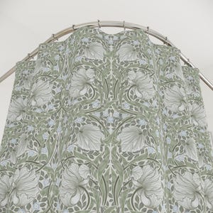 May include: A shower curtain featuring a repeating botanical pattern with large white flowers, sage green foliage, and small light blue blossoms. The fabric hangs from a curved metal rod with silver rings, displaying a symmetrical floral design.