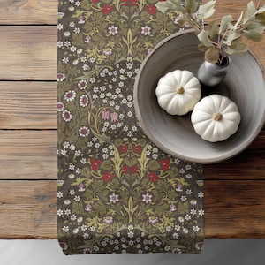 May include: A table runner featuring a dark brown background with a repeating botanical pattern of white, red, and light green flowers and leaves. A gray ceramic bowl sits on the runner, containing two white pumpkins and a dried plant stem.