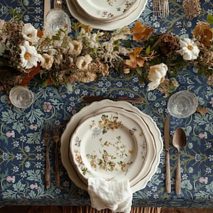 May include: A beautifully set table with floral patterned plates, silverware, and a white linen napkin. The table is adorned with a blue floral tablecloth and a centerpiece of flowers and foliage. The plates have a gold trim.