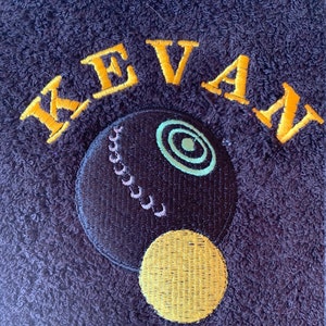 May include: A dark purple towel with the name "KEVAN" embroidered in yellow. A black and white bowling ball with a yellow circle is embroidered below the name.