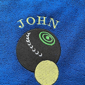 May include: A blue towel with a black and white bowling ball and a yellow bowling ball embroidered on it. The name "JOHN" is embroidered in yellow above the bowling balls.