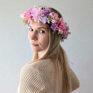 May include: A floral crown featuring a variety of pink, purple, and white flowers. The crown is composed of large, textured blooms and smaller blossoms, creating a vibrant and colorful accessory. The flowers are arranged on a thin, flexible base.