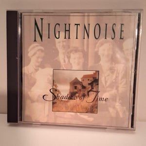 May include: A Nightnoise CD case with the album title "Shadow of Time." The cover features a sepia-toned image of people and a small landscape scene with a building. The band name "Nightnoise" is printed at the top.