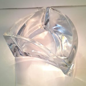 May include: A clear, sculpted glass ashtray with a unique, geometric design. The ashtray has a curved, open shape with multiple facets, creating a play of light and shadow. The glass is transparent, allowing a view through the object.