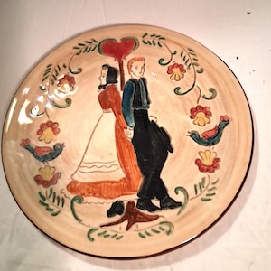 May include: A round, hand-painted ceramic plate with a folk art design. The plate features two figures in traditional clothing, a red heart, and floral and bird motifs in shades of red, green, and blue on a cream background.