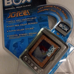 May include: A silver Juice Box personal media player in its packaging. The player has a small screen and buttons for controlling music and video playback. The packaging is blue and features the text "Turn on the Juice!" and "MP3 & JPEG Compatible!"