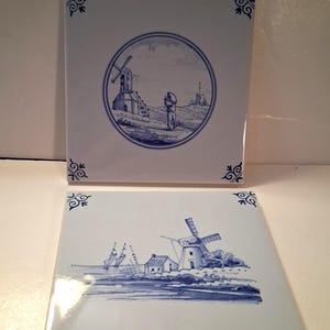 May include: Two square ceramic tiles with blue and white windmill designs. One tile features a circular scene with a windmill and a person. The other tile shows a landscape with windmills and buildings. Decorative corner accents.