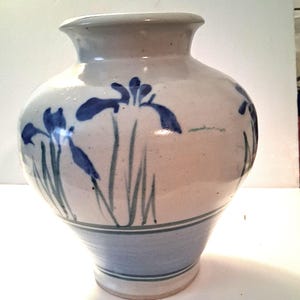 May include: A ceramic vase with a cream-colored glaze featuring hand-painted blue iris flowers and green stems. The base of the vase is decorated with horizontal blue bands, and the vessel has a wide, rounded body with a flared rim opening.