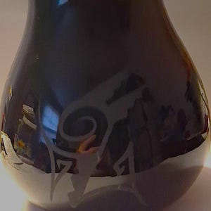 May include: A glossy black glass vase with a rounded, bulbous body. The surface features a light-colored, etched graphic design consisting of a central spiral element and angular, geometric shapes extending downward toward the base of the vessel.