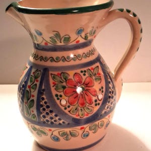 May include: A hand-painted ceramic pitcher with a cream-colored base. The pitcher features a floral design with red, blue, and green accents. The handle and rim are decorated with green and blue details. The pitcher is approximately 6 inches tall.