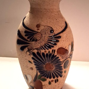 May include: A tan ceramic vase with a textured surface, featuring a detailed design of a bird in flight and a large blue flower. The vase has a narrow neck and a rounded body, with brown and white accents.