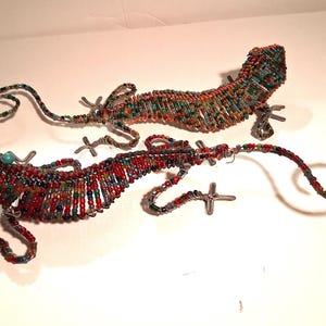 May include: Two beaded lizard sculptures. The lizards are made of colorful beads in shades of red, green, blue, and orange, with silver wire legs and tails. One lizard has turquoise beads for eyes.