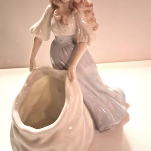 May include: A porcelain figurine of a woman in a flowing gown, holding a large, open sack. The woman has long, wavy hair and is dressed in a white blouse and a pale blue dress. The figurine is a decorative item.