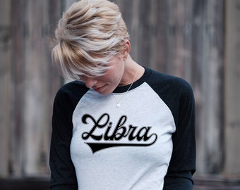 Puede incluir: Una camiseta de estilo béisbol blanca y negra con la palabra "Libra" en una fuente de letra negra en la parte delantera.