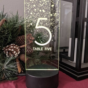 May include: A clear acrylic table number with a white number 5 and the text "TABLE FIVE" on a black base with a white dot pattern. The base is lit with a warm white light.