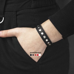 Könnte beinhalten: Schwarzes Lederarmband mit silbernen Nieten. Das Armband wird an einem Handgelenk getragen.