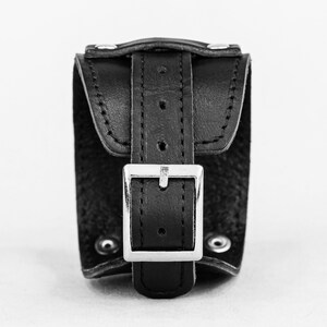 May include: A black leather wrist cuff with a silver-colored buckle. The cuff has a rectangular shape with a flap that fastens with a buckle. The cuff is secured with rivets. The background is plain white.