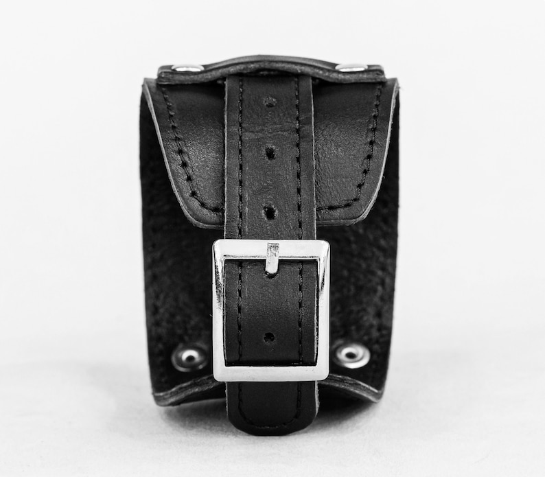 May include: A black leather wrist cuff with a silver-colored buckle. The cuff has a rectangular shape with a flap that fastens with a buckle. The cuff is secured with rivets. The background is plain white.