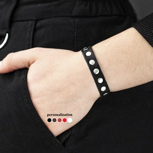 Puede incluir: Pulsera de cuero negro con tachuelas plateadas. La pulsera es ajustable y tiene un diseño sencillo.
