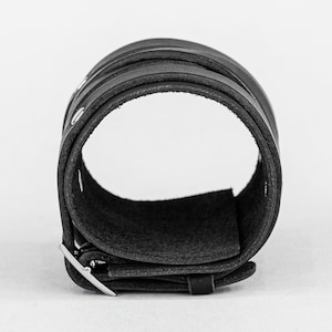 May include: A black leather cuff bracelet with a silver buckle. The bracelet is made of multiple layers of leather and has a smooth, polished finish.