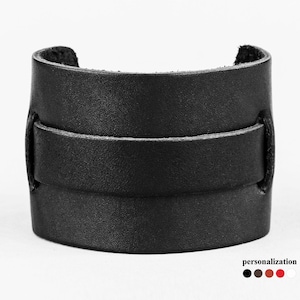 以下が含まれることがあります： 前面にシングルストラップが付いたブラックレザーのカフブレスレット。ブレスレットはさまざまな色からお選びいただけます。