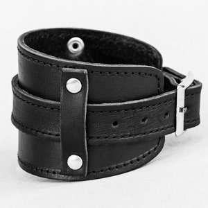 May include: A black leather wrist cuff with a wide band and a buckle closure. The cuff features two smaller straps and is secured with silver-toned rivets and a buckle. The cuff is designed to wrap around the wrist.