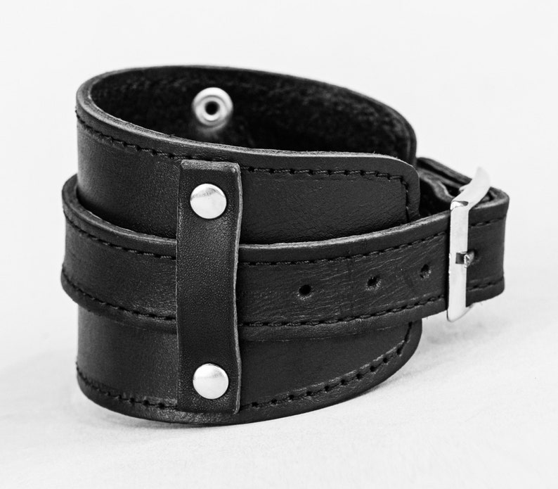 May include: A black leather wrist cuff with a wide band and a buckle closure. The cuff features two smaller straps and is secured with silver-toned rivets and a buckle. The cuff is designed to wrap around the wrist.