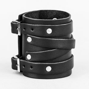 May include: A black leather cuff bracelet with silver metal buckles and studs. The bracelet is made of multiple layers of leather that are wrapped around the wrist.