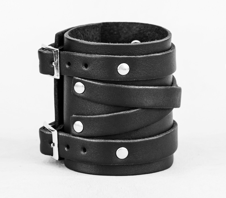 May include: A black leather cuff bracelet with silver metal buckles and studs. The bracelet is made of multiple layers of leather that are wrapped around the wrist.