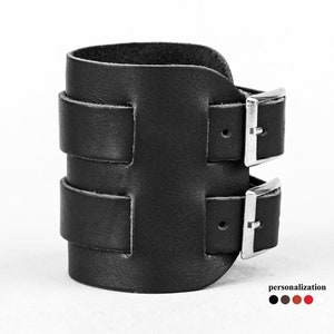 May include: Black leather cuff bracelet with two silver buckles. The bracelet is adjustable and has a simple, modern design.