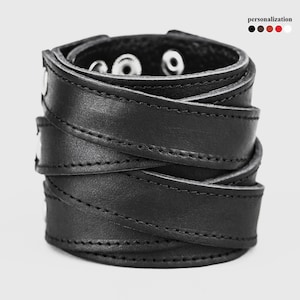 May include: Black leather cuff bracelet with a wrap-around design. The bracelet features a series of overlapping straps secured with silver-tone snaps. The edges are stitched with black thread. The word "personalization" is in the upper right corner.