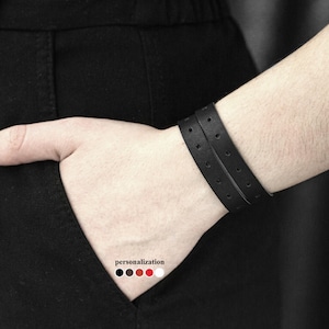 May include: A black leather double wrap bracelet with a row of small holes on each strap. The bracelet is shown on a person's wrist.